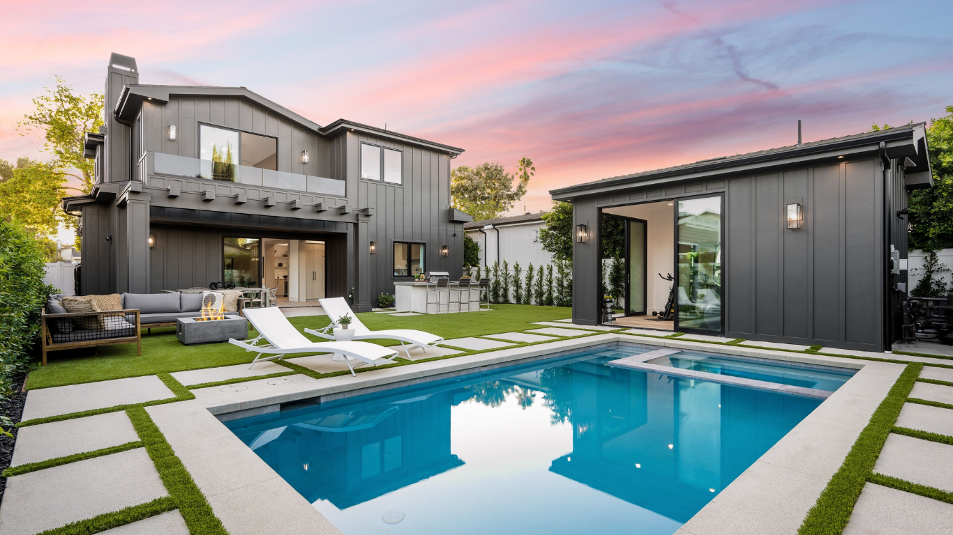 Modern house with pool, yard, and detached structure. Dark gray exterior, blue pool, green grass, and pink/orange sky.