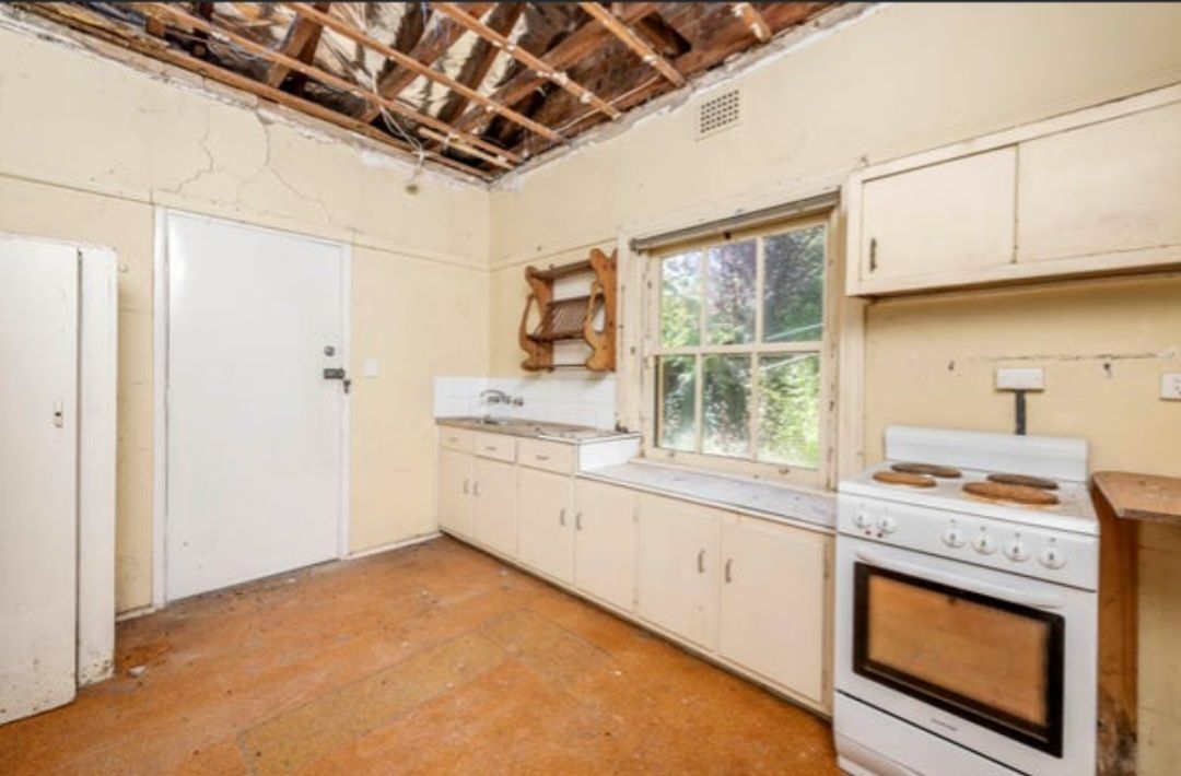 Old, Worn Kitchen With Exposed Ceiling. White Cabinets, Stove, and Door. Window and Sink Along One Wall — Elimar Construction in Goulburn, NSW