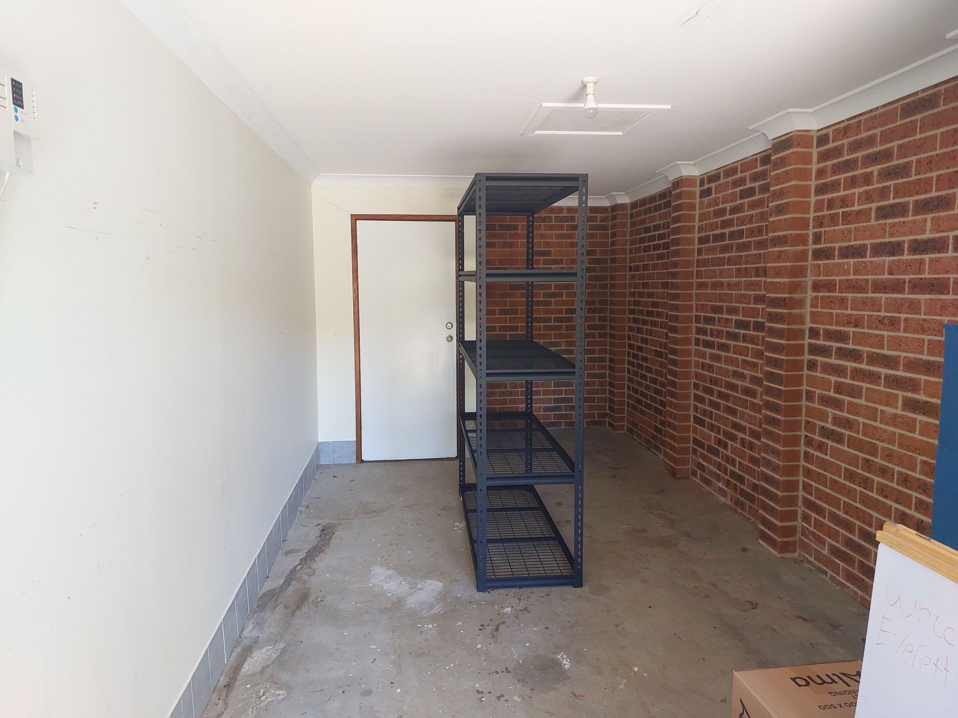 Empty Garage With Brick Wall, White Wall, Metal Shelving, and a Door. Concrete Floor and Overhead Lights — Elimar Construction in Goulburn, NSW