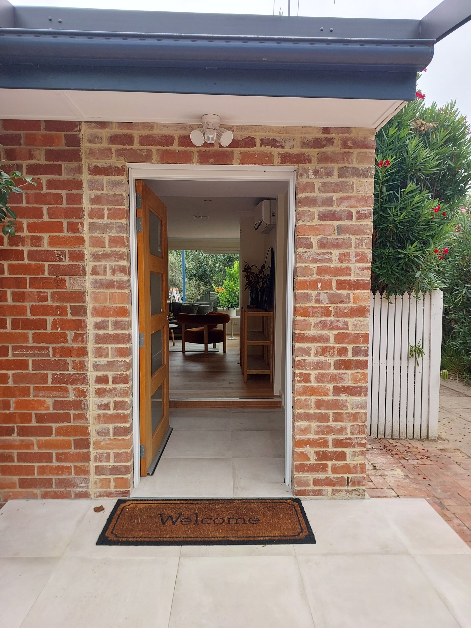Brick doorway with open wooden door, doormat, view of interior with furniture and greenery -Elimar Construction in Goulburn, NSW
