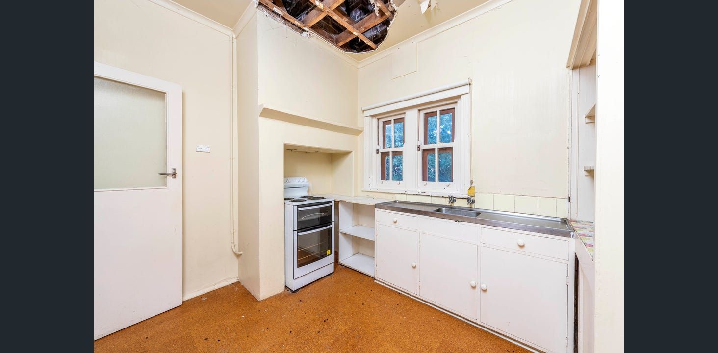 Small, Dated Kitchen With White Cabinets, a Stove, and a Window. the Ceiling is Damaged — Elimar Construction in Goulburn, NSW