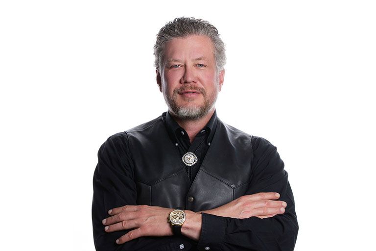 Man with arms crossed, wearing a black shirt and vest, smiling with a watch and a silver bolo tie.