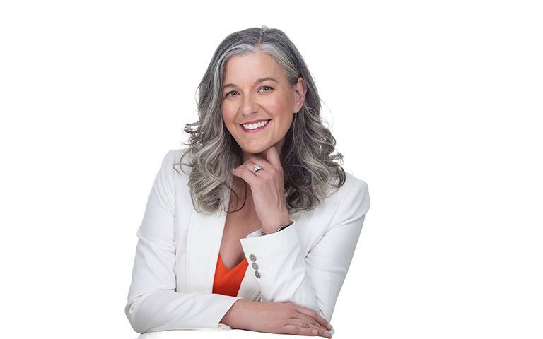 Woman in white blazer, orange top, silver hair, smiles, hand to face, white background.