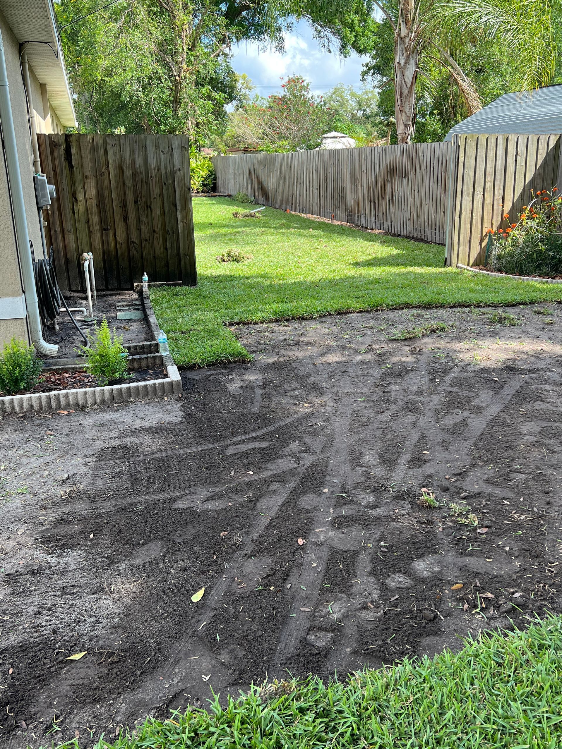 Yard with a wooden fence, grass, and a dark-colored, uneven surface.