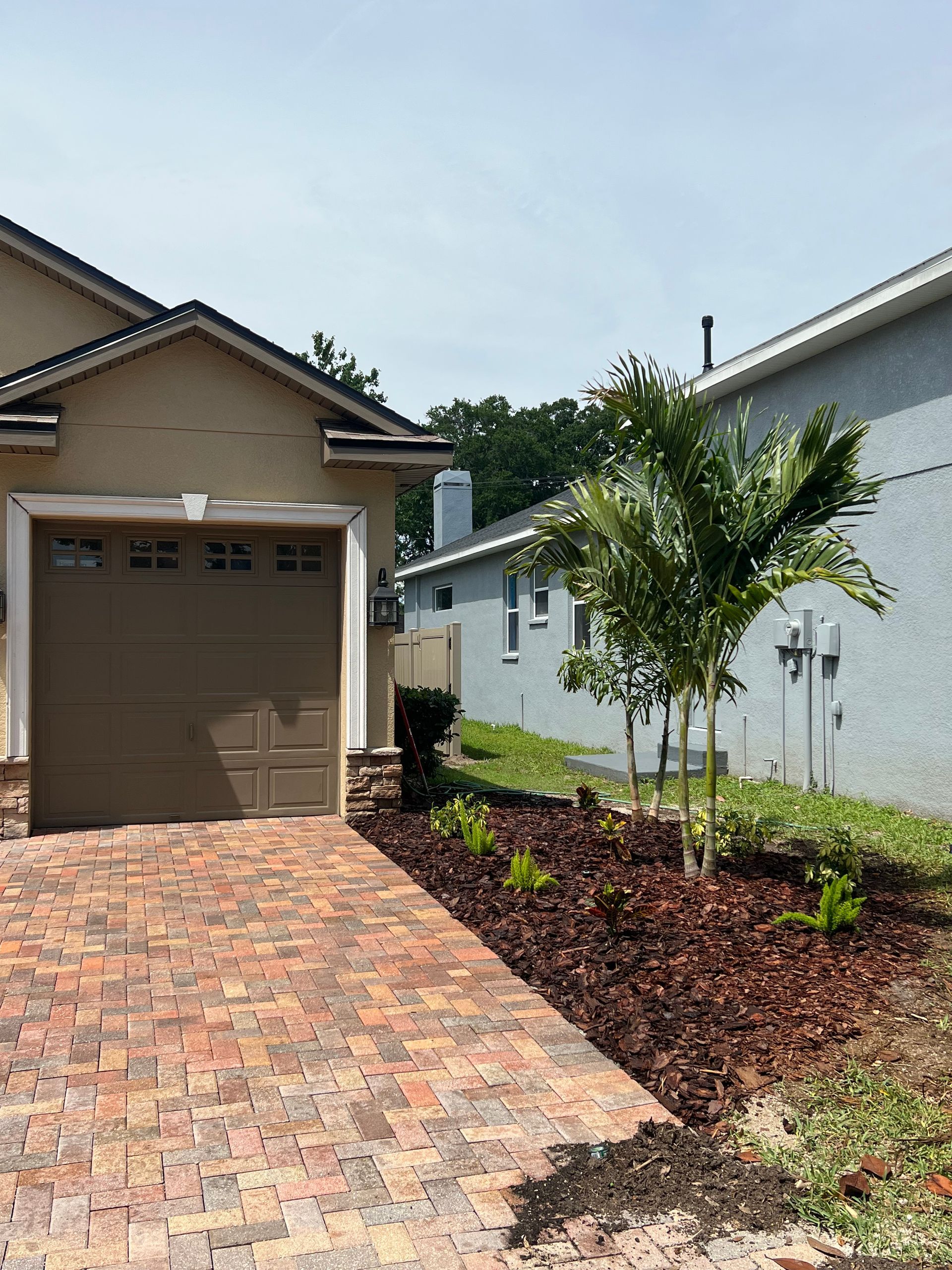 Driveway with brick pavers, garage, and landscaping with a palm tree, sunny day.