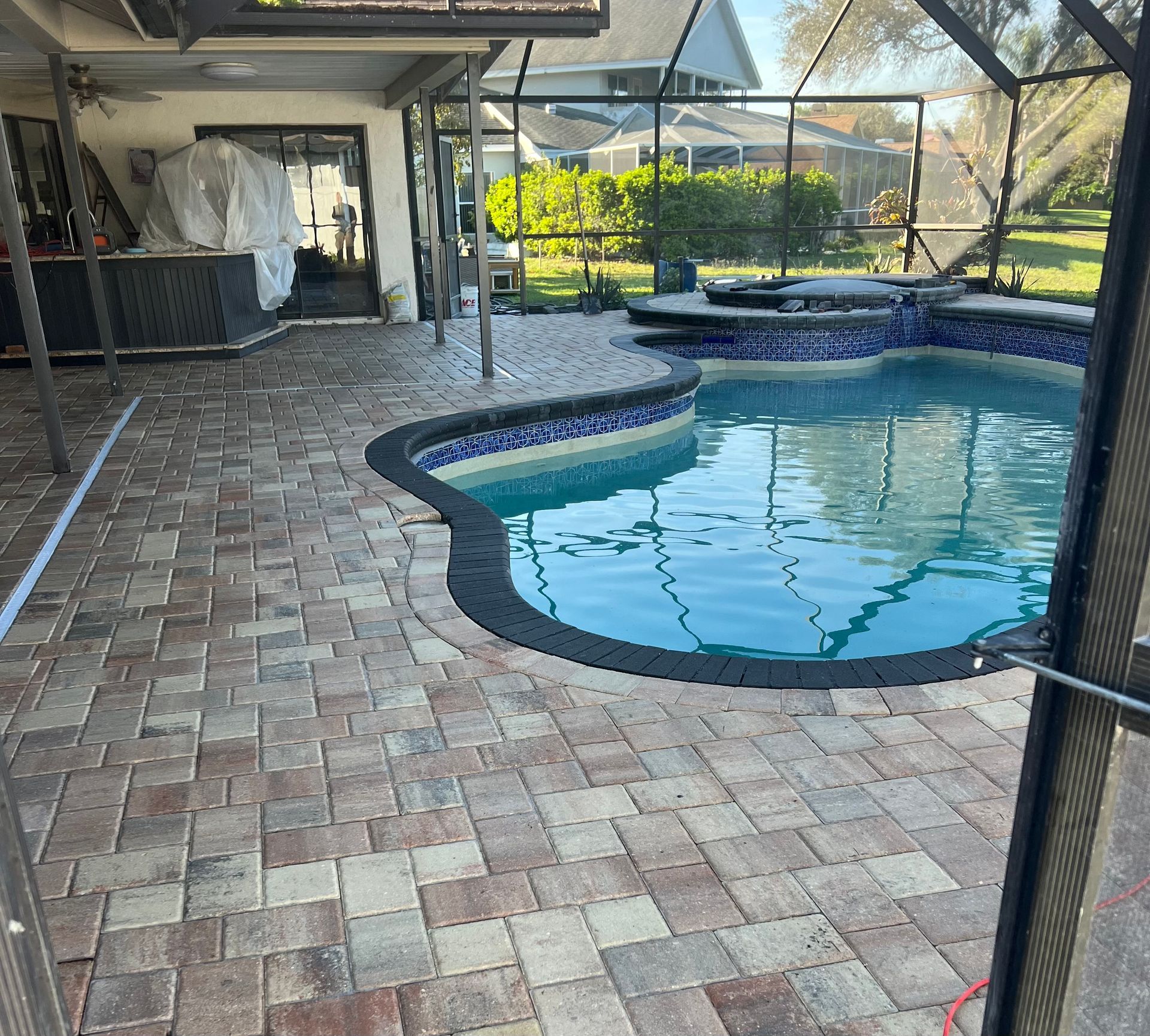 Brick patio with a pool, enclosed by a screen. Blue pool with mosaic tiles, and green grass in the background.
