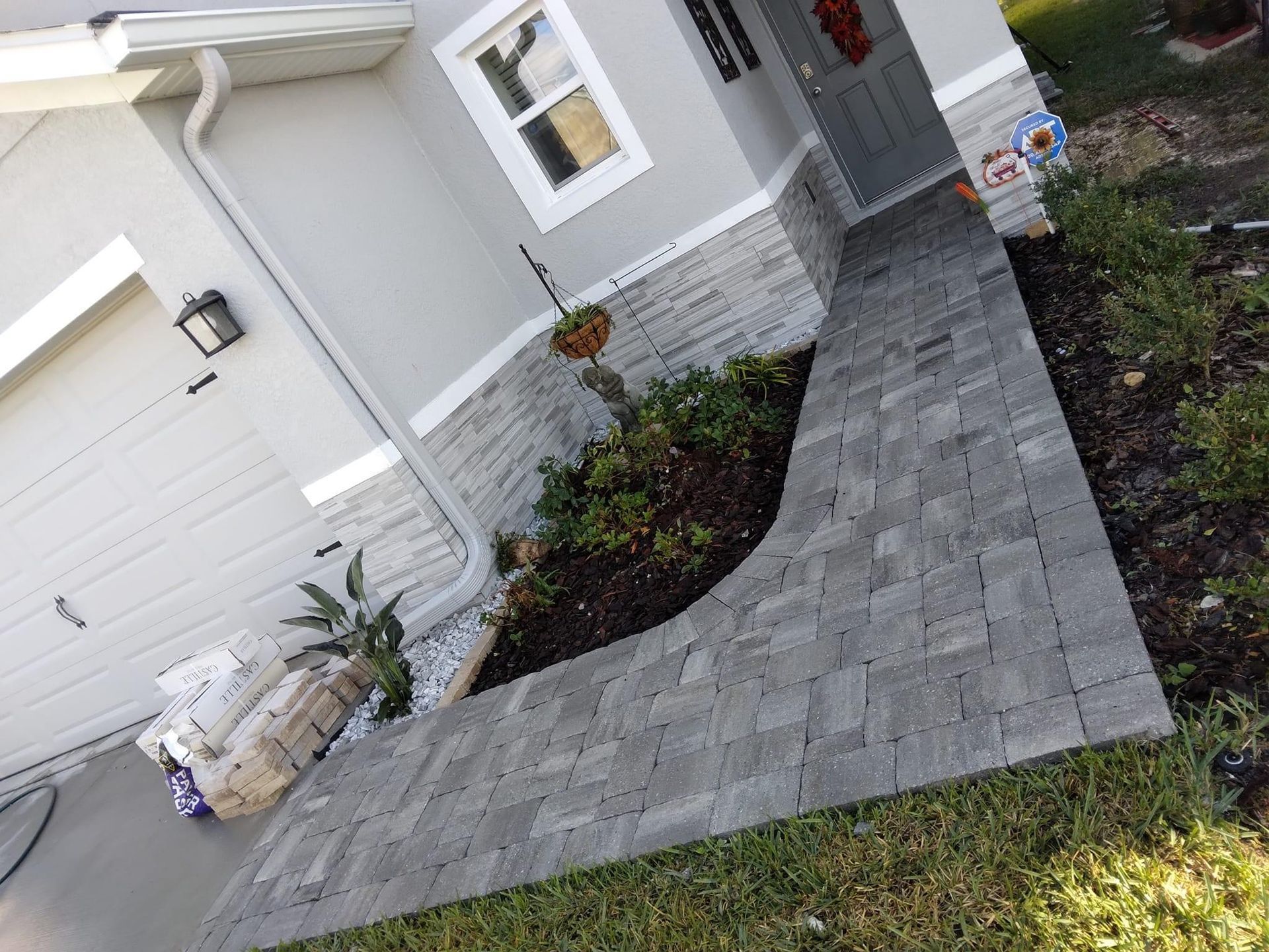 Grey brick walkway curves to a house entrance with stone facade, next to a small garden.
