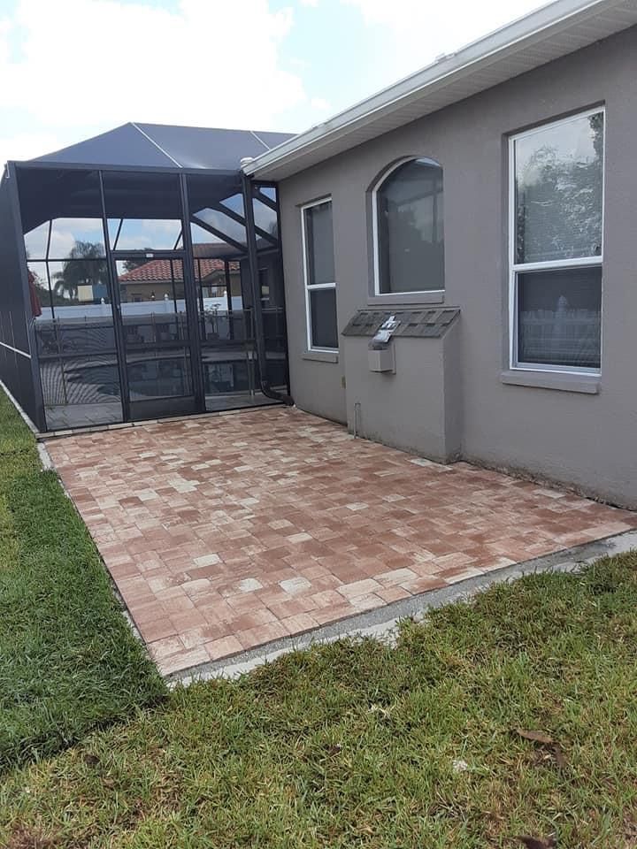 Brick patio beside a screened pool enclosure and a gray house with windows. Green grass surrounds.
