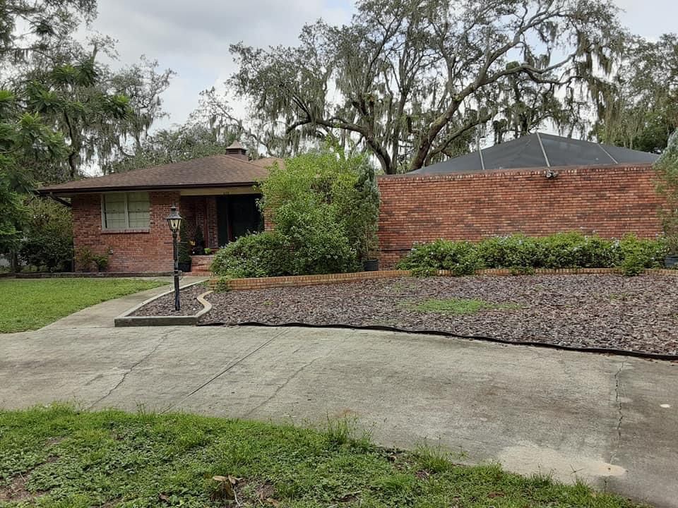 Brick house with dark roof and brick wall in front yard, with driveway and trees in the background.