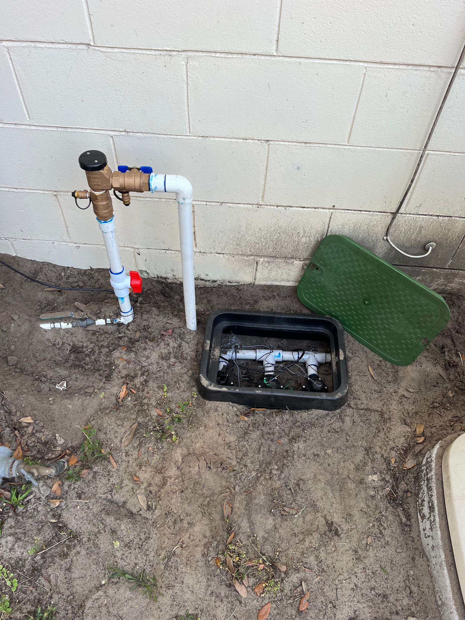 Sprinkler system components near a white brick wall, including a valve, pipes, and an open irrigation control box.