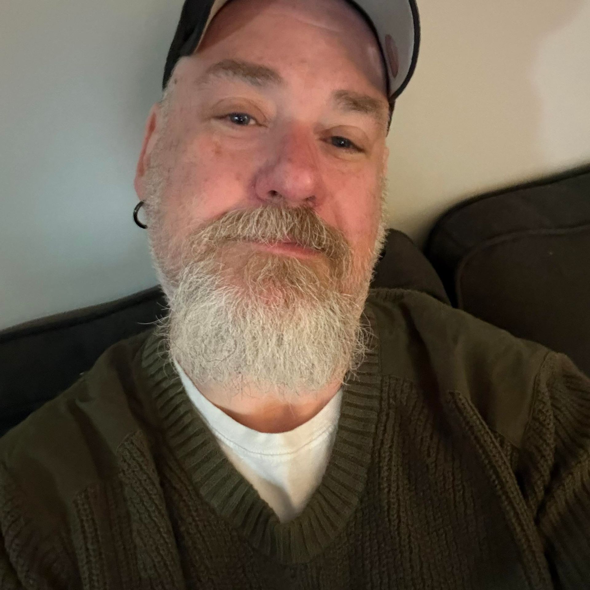 Man with a beard and baseball cap sits on a couch, looking at the camera.