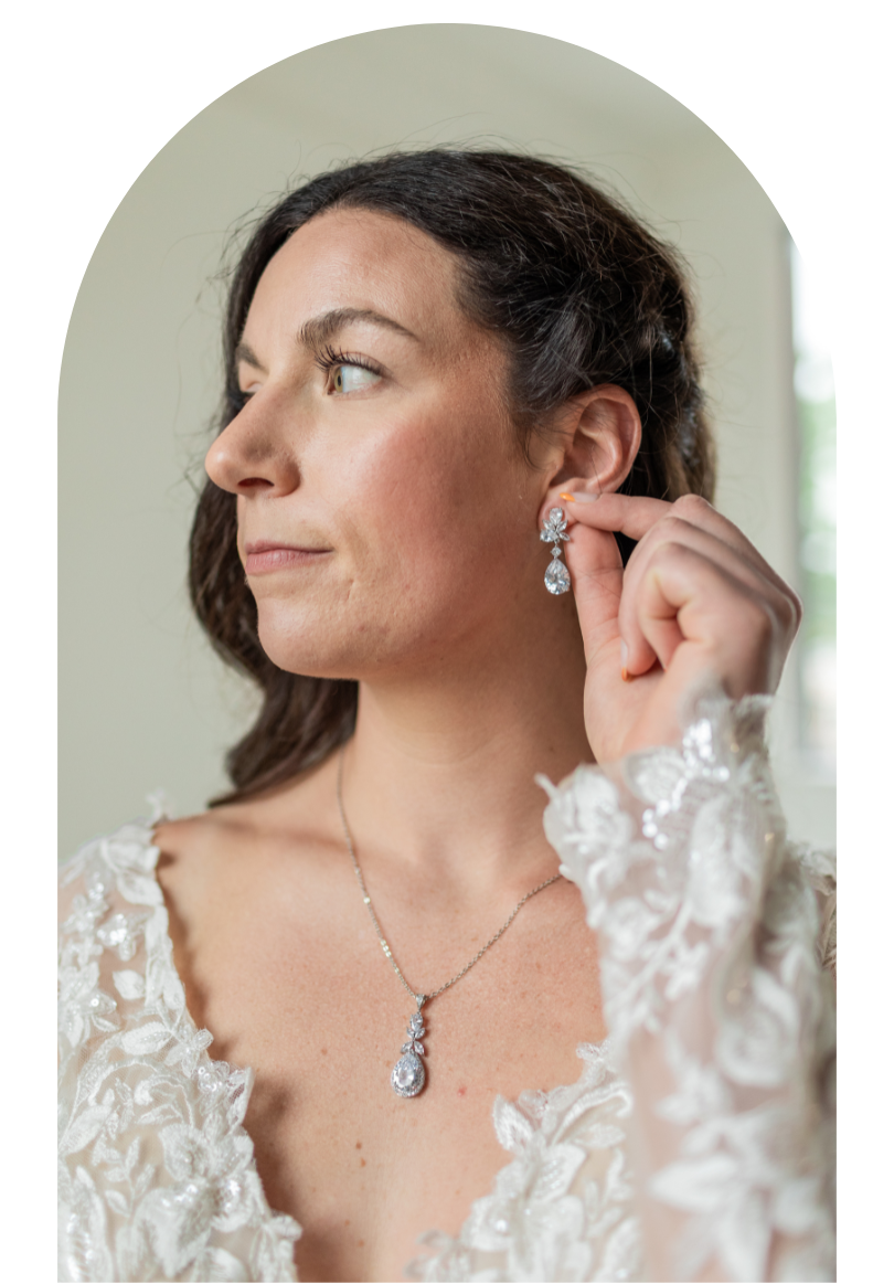 Woman in a white lace dress adjusting a dangling earring, wearing a matching necklace against a light background