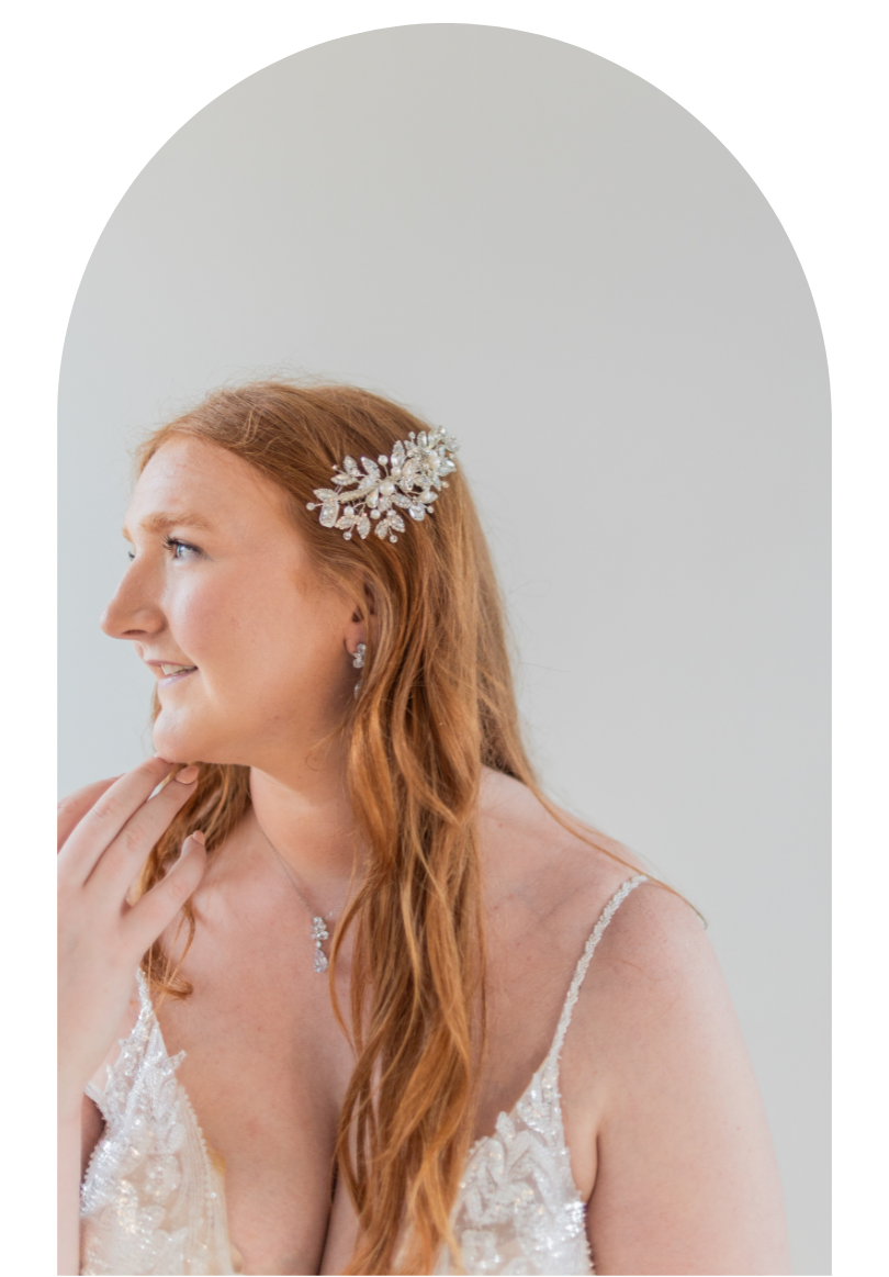 Woman in a white dress with floral hairpiece, looking left against a plain white arched background