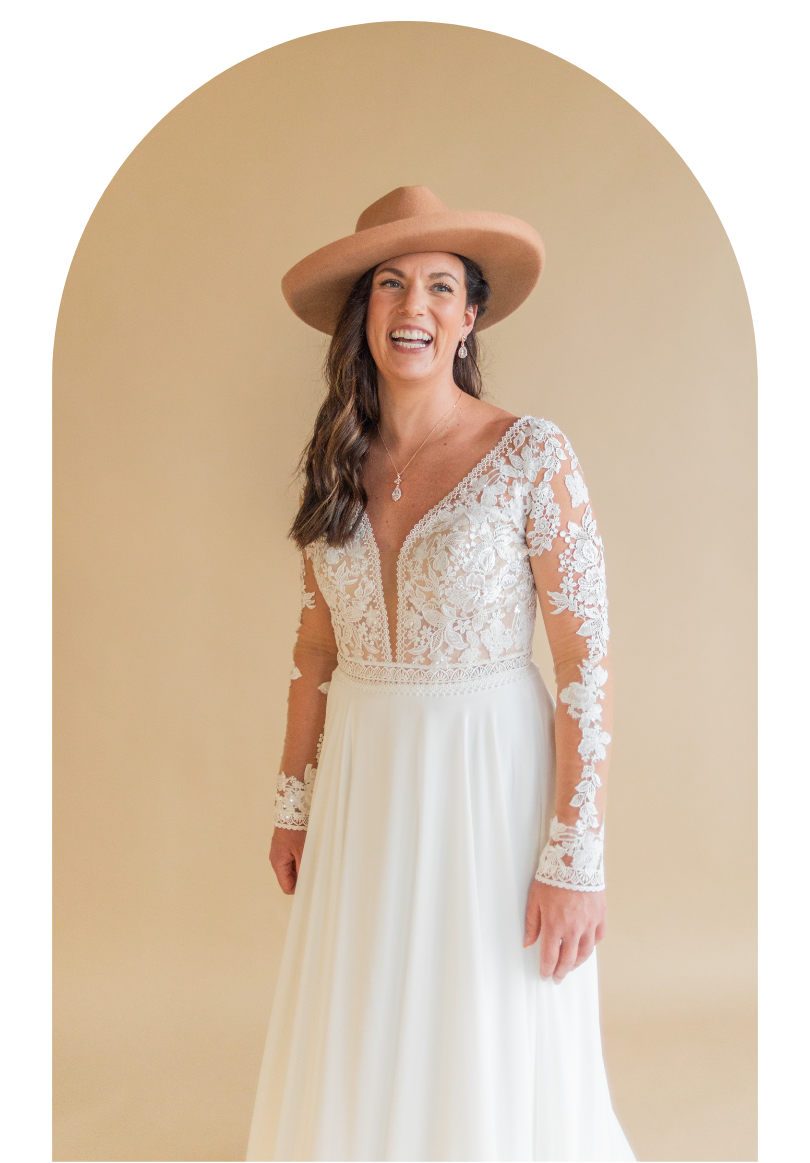Woman in a white lace wedding dress and tan hat standing in an arched doorway, smiling.