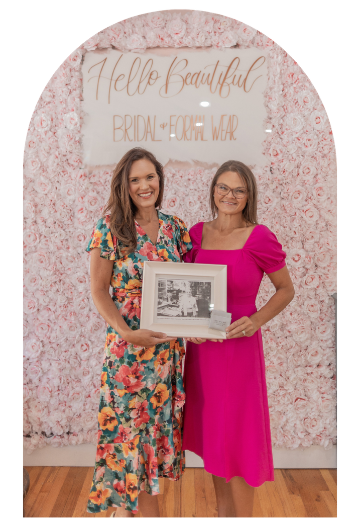 Two women pose holding a framed item in front of a pink floral wall backdrop.