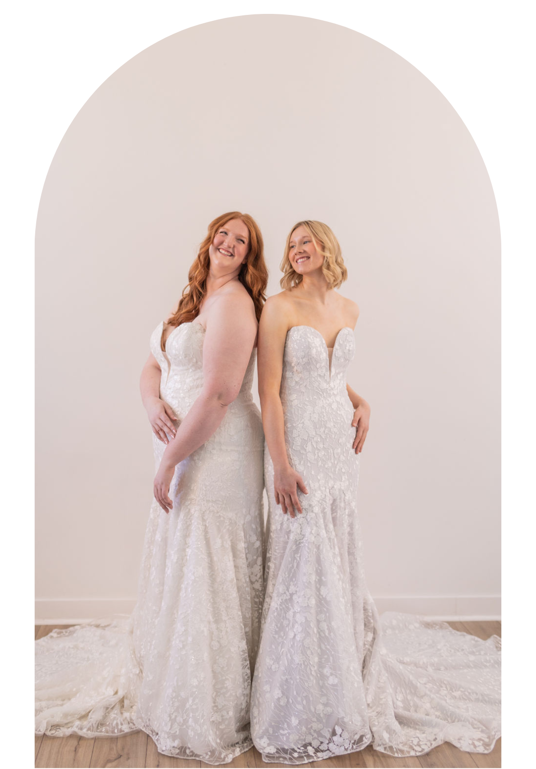 Two women in white lace gowns posing in a studio against a soft arched backdrop.