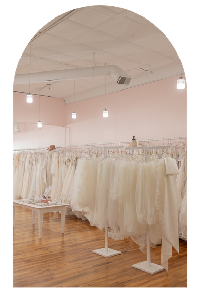 Rows of cream dresses on racks in a pastel pink boutique with arched ceiling and wood floor