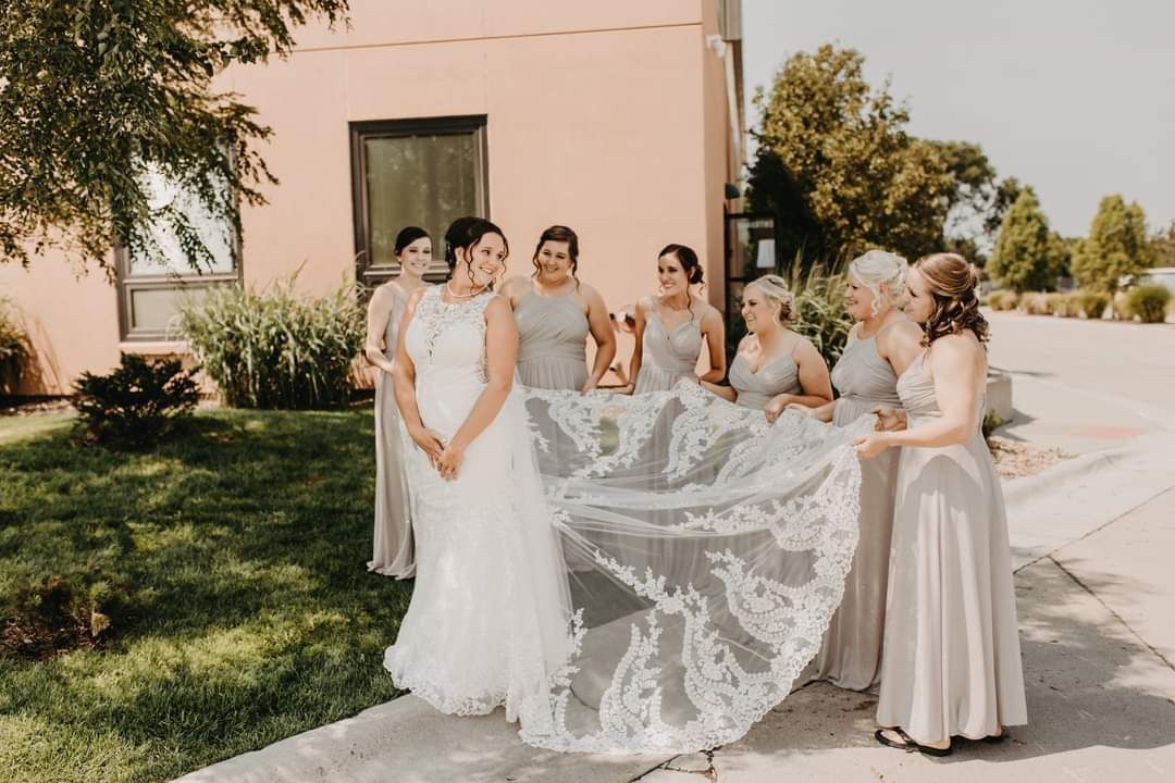 Bridesmaids gather around a bride in a white gown outdoors beside a pink building.