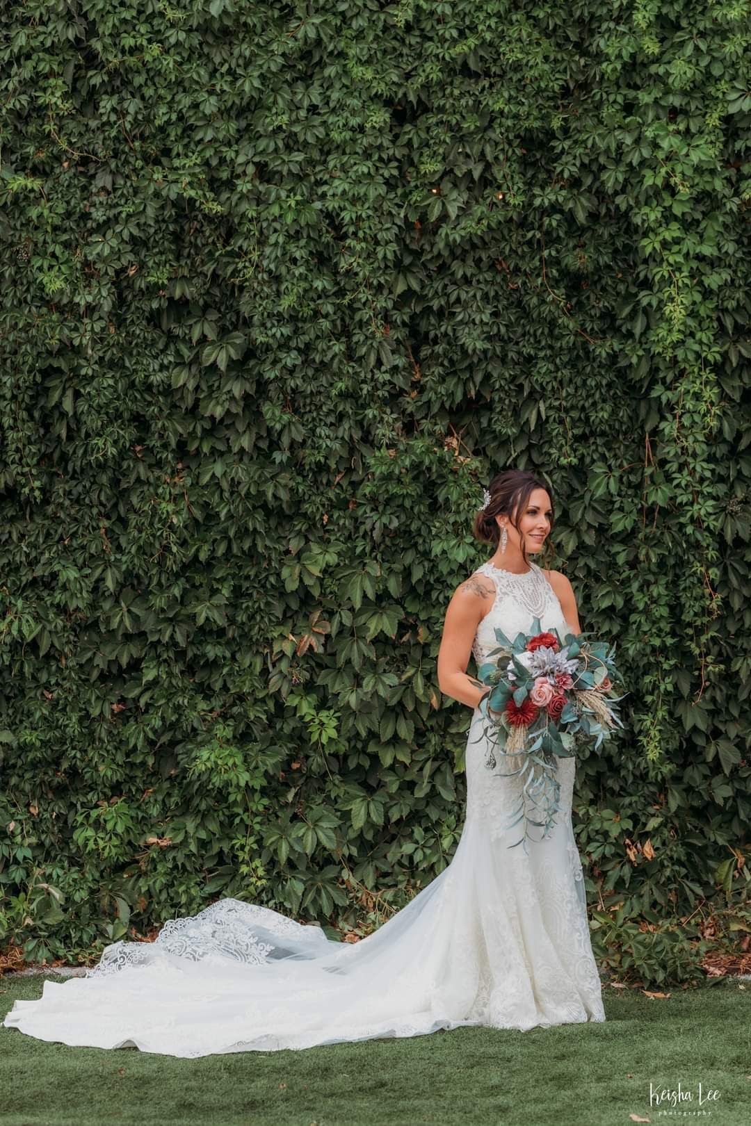 Bride in a white lace gown holding a colorful bouquet before a lush green ivy wall