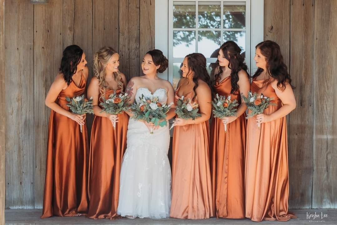 Bride with five bridesmaids in peach dresses standing against a wooden wall, holding bouquets and smiling