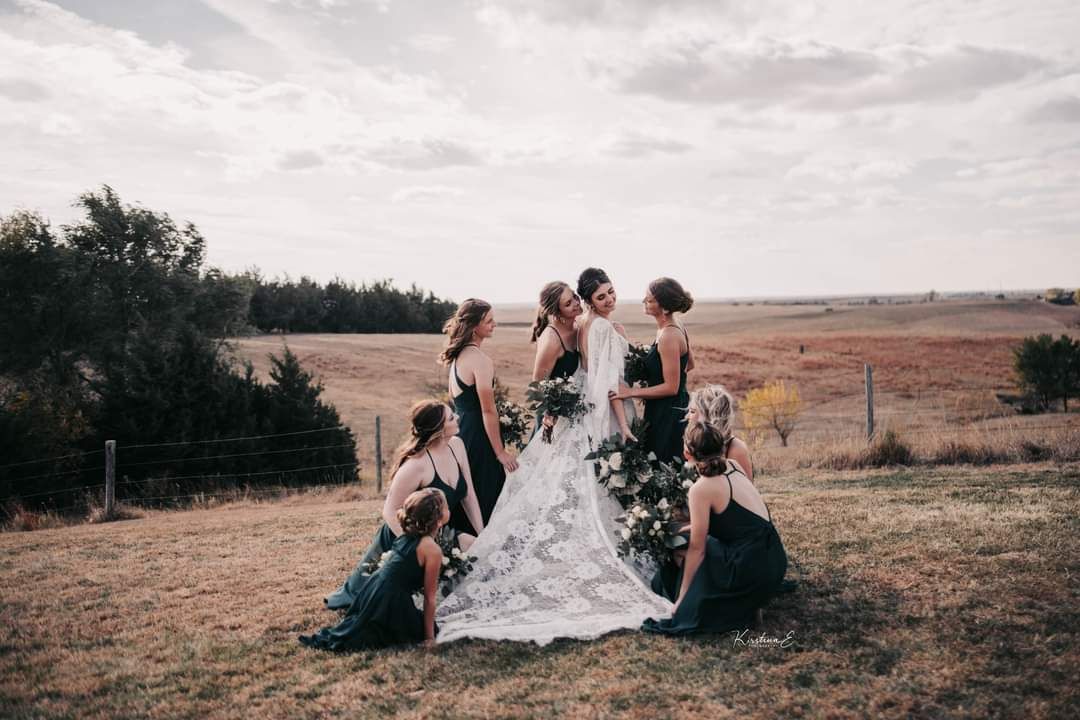 Wedding party posing in a field, with the bride in a white dress surrounded by attendants in muted tones.