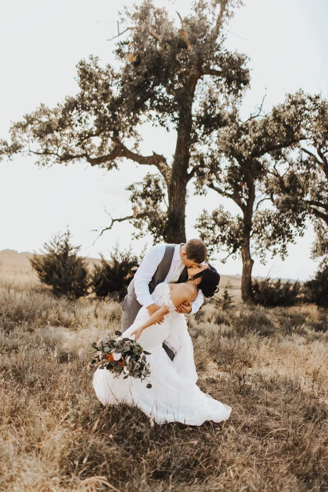Couple in white wedding attire embracing in a dry field beneath a large tree