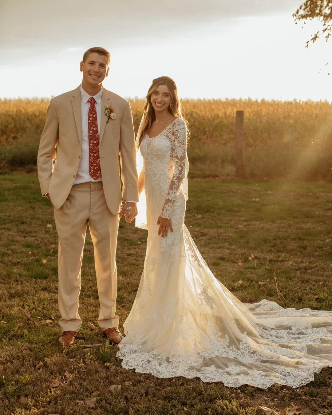 Bride and groom standing in a field at sunset, holding hands, with the bride’s long lace train trailing behind.