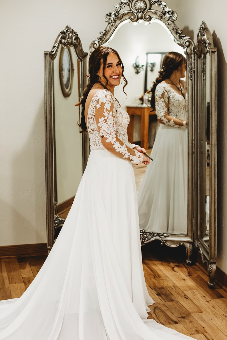 Woman in a white lace gown standing by an ornate mirror, smiling in a bridal shop.