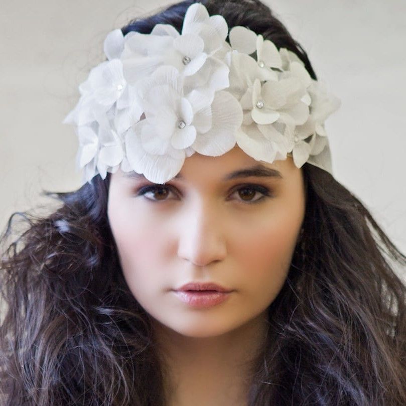 Woman with dark curly hair wearing a white floral headband, facing forward against a light background