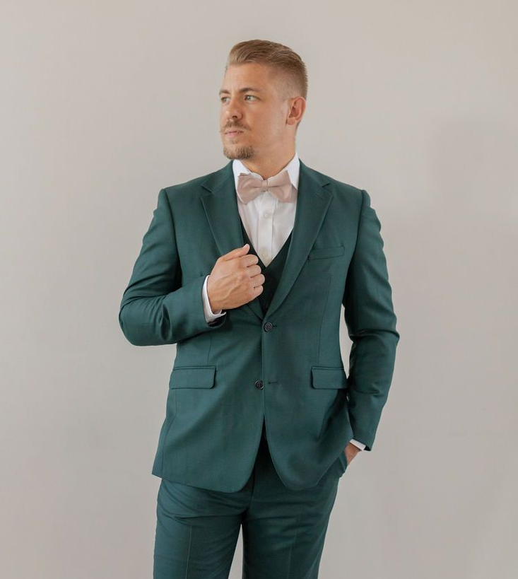Man in a teal suit and white shirt standing against a plain wall, adjusting his jacket and looking aside