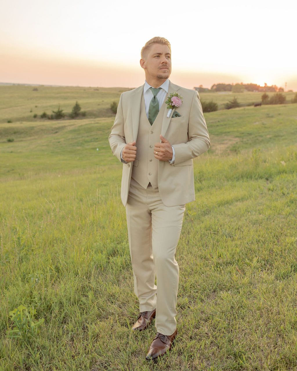 Man in a light beige suit holding a boutonniere, standing in a grassy field at sunset.