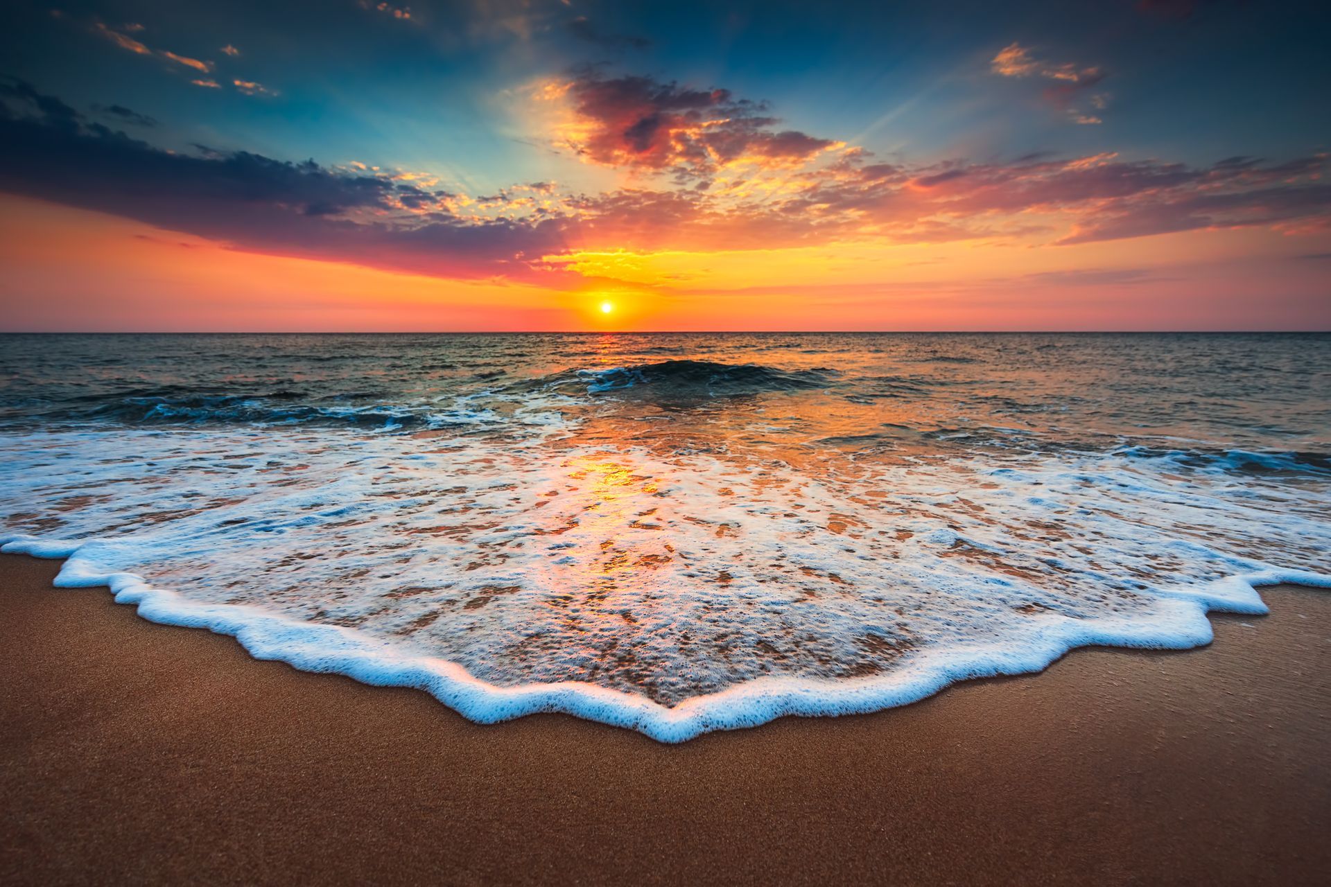 Sunset over the ocean, with waves crashing on the shore. Orange, yellow and purple hues in the sky.