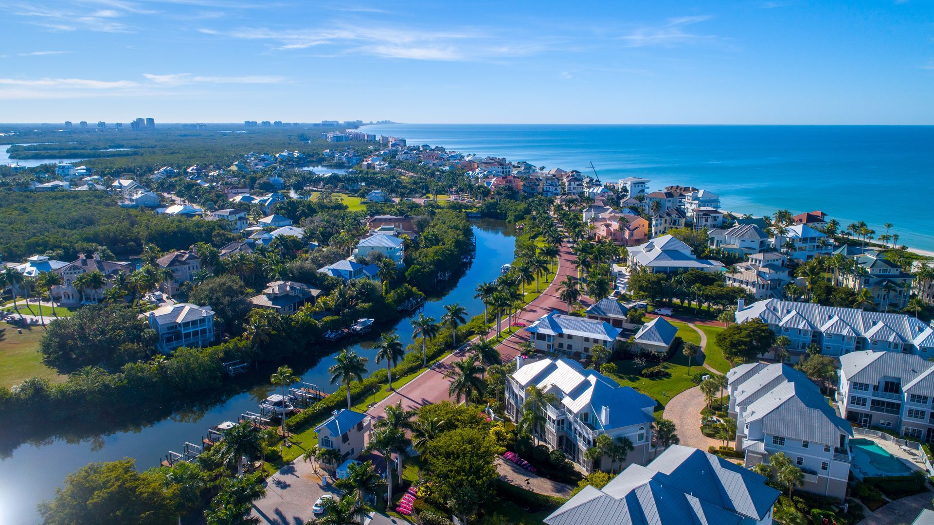 Aerial view of a coastal neighborhood with houses, a narrow canal, lush greenery, and the ocean under a clear blue sky.
