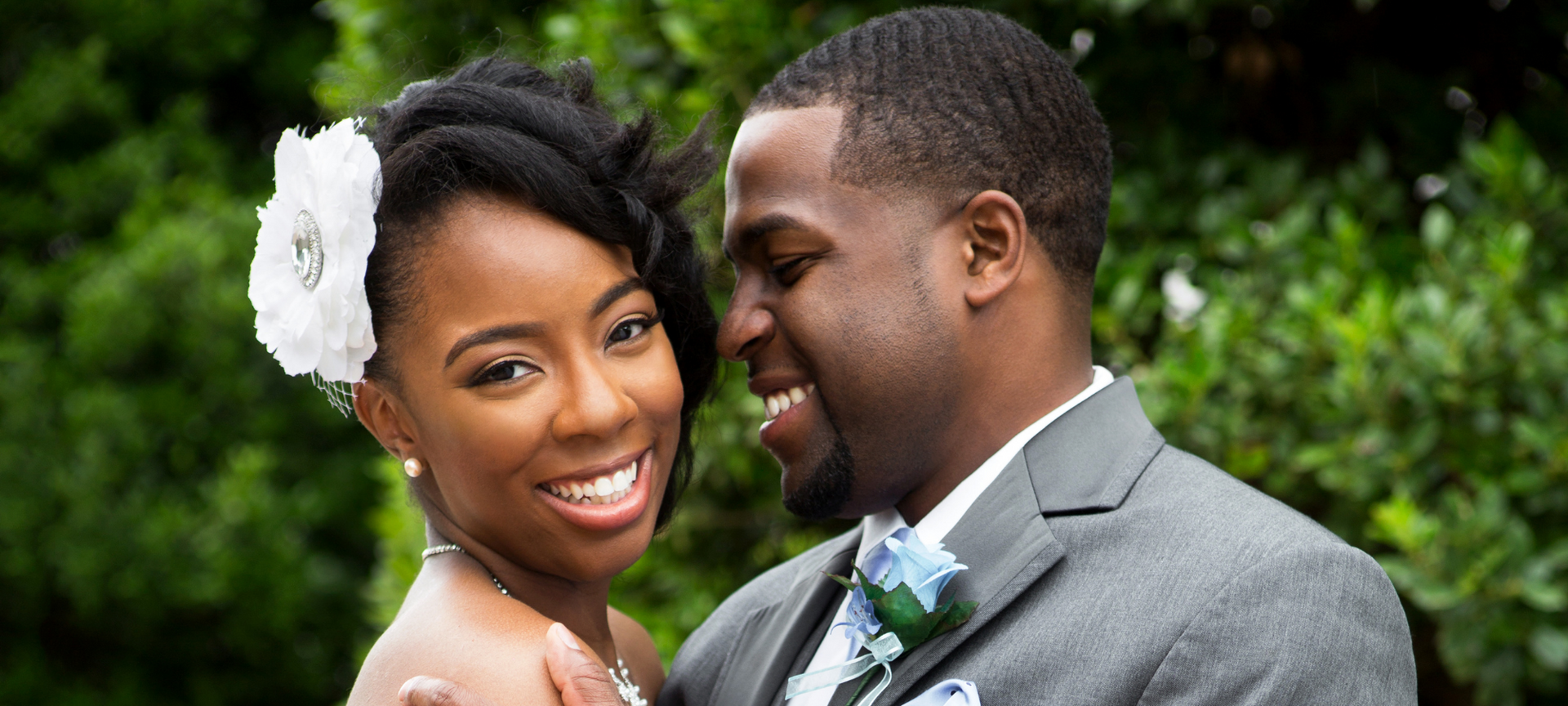 Smiling couple in formal attire outdoors, with greenery in the background.