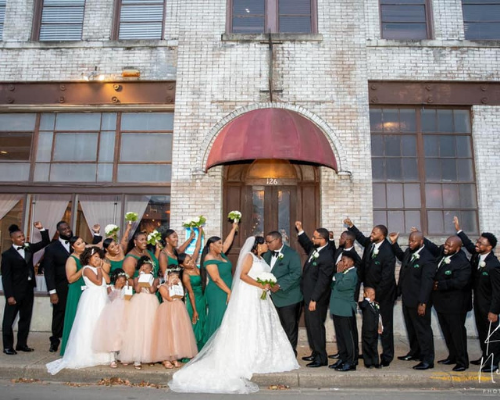 Wedding party posing outside a brick building, with bride in white and attendants in green and black attire.