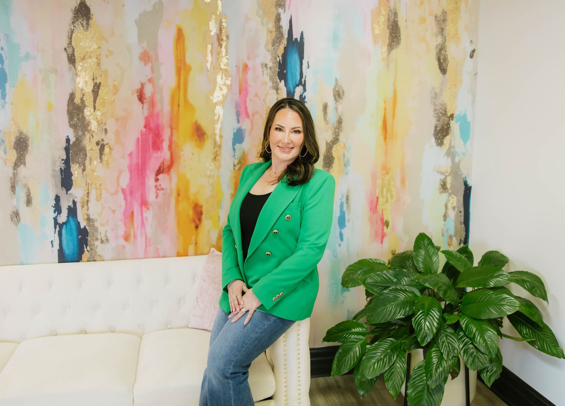 Woman in a green blazer sitting on a white couch beside a leafy plant and colorful abstract wall art.