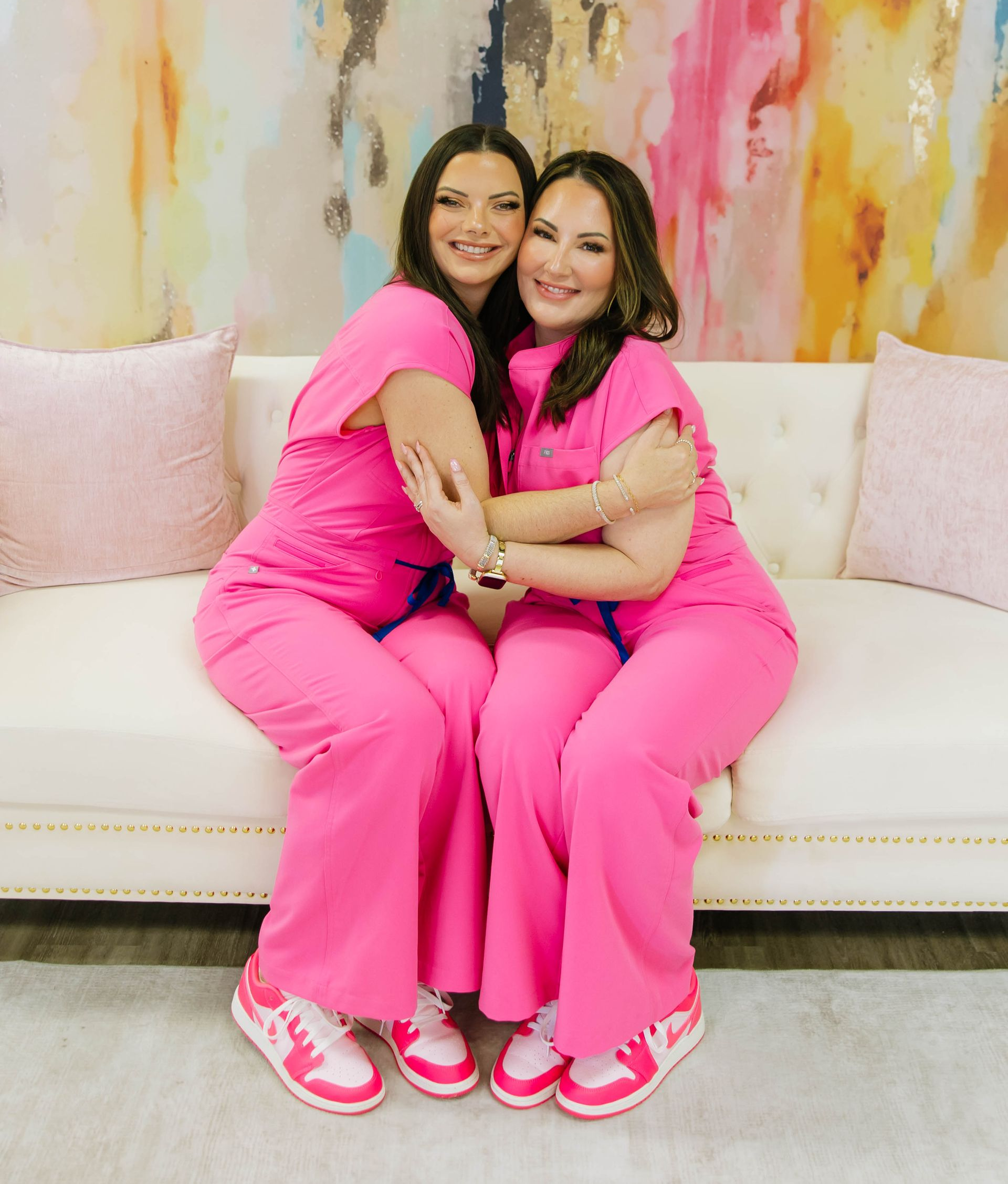 Two people in bright pink scrubs sitting on a white couch, smiling and embracing.