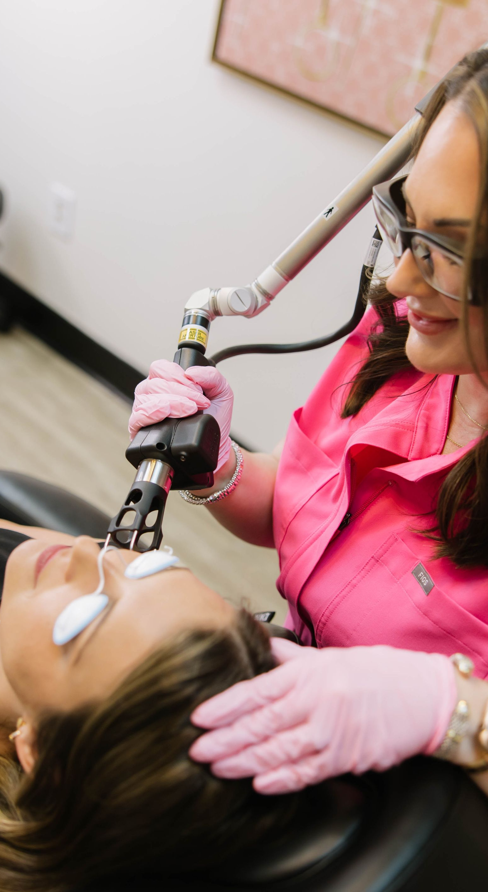 Technician using a laser device on a patient’s face in a pink-gloved treatment room