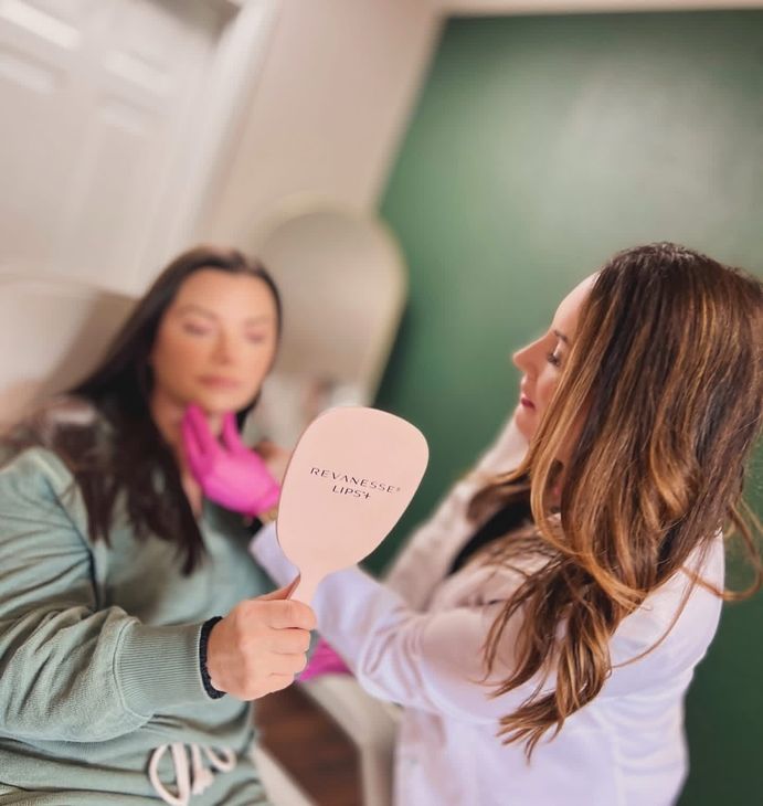 A woman showing another woman her reflection in a hand held mirror.