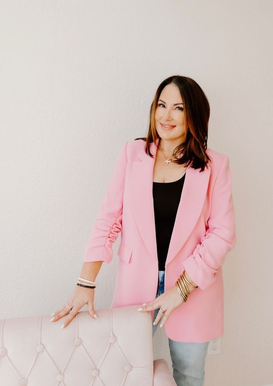 Woman in a pink blazer standing beside a tufted chair against a white wall.