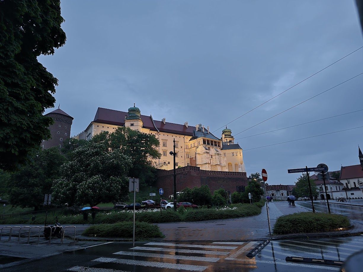Lit castle on a hill at dusk beside a wet path and trees under a cloudy sky