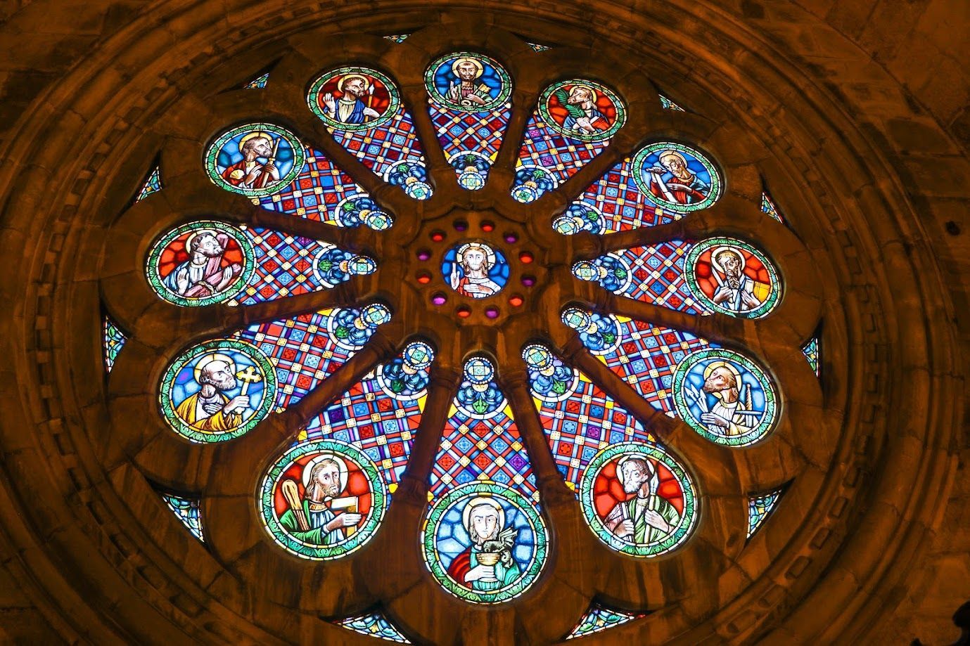 Circular stained-glass rose window with colorful religious scenes in a stone cathedral ceiling