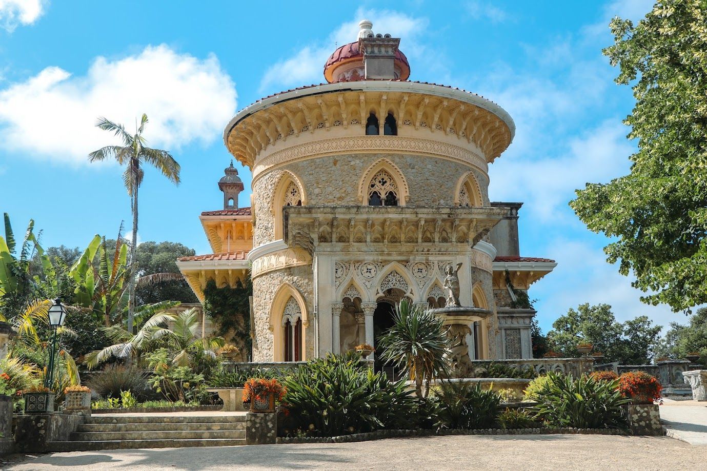 Ornate historic villa with a round tower, surrounded by lush greenery under a bright blue sky.