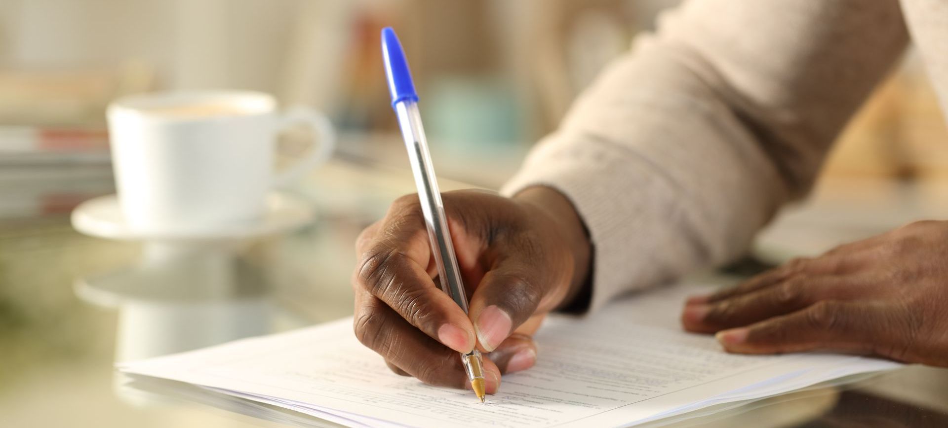 Hand writing on a notebook with a blue pen beside a cup on a table