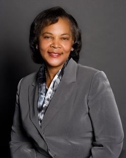 Studio portrait of a person in a gray blazer, smiling against a dark background