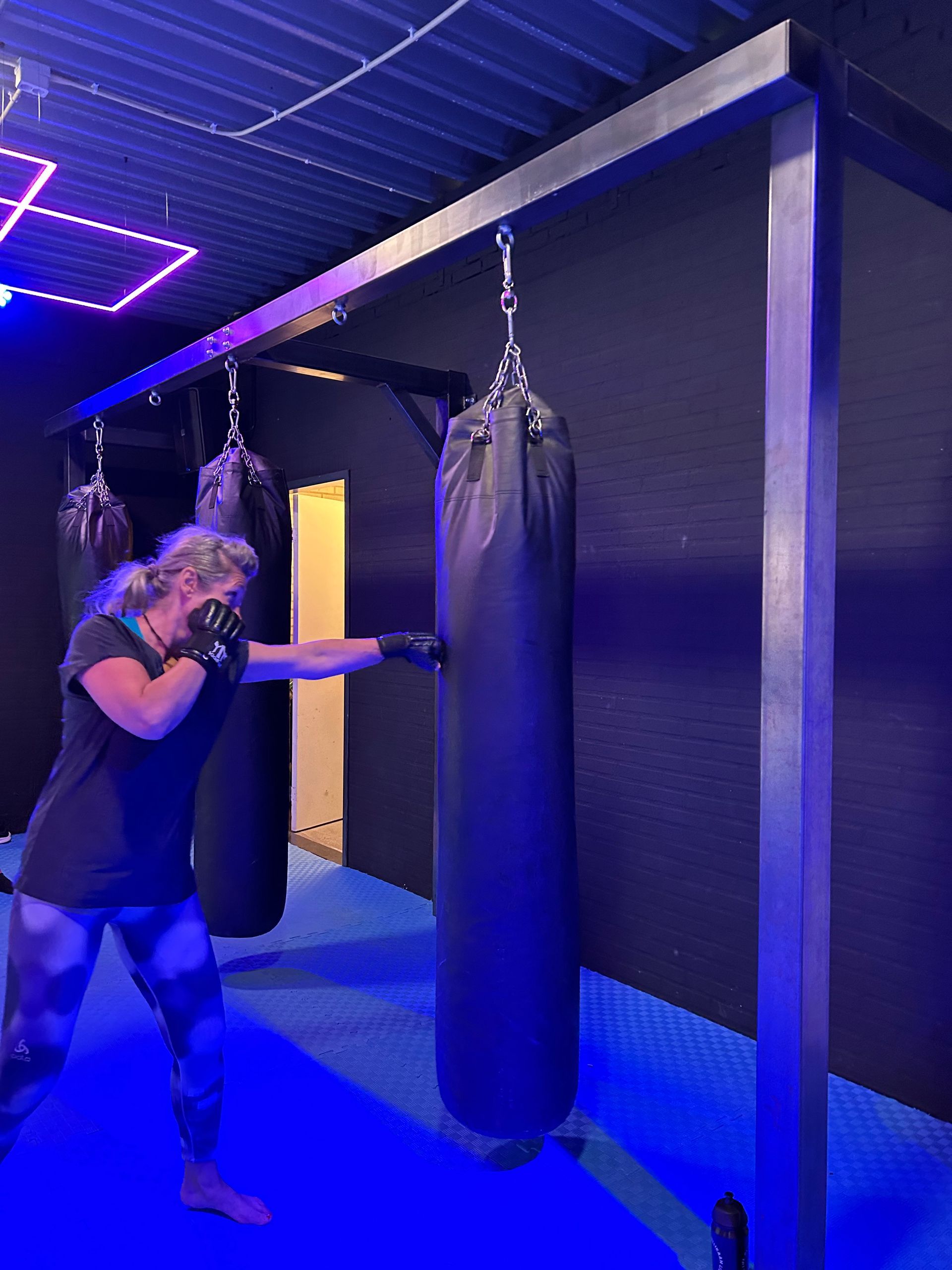 Woman punching a black heavy bag in a boxing gym with blue and purple lighting.
