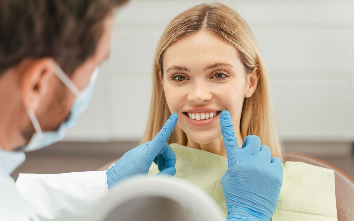 Dentist in mask and gloves points at woman's teeth, smiles, in office.