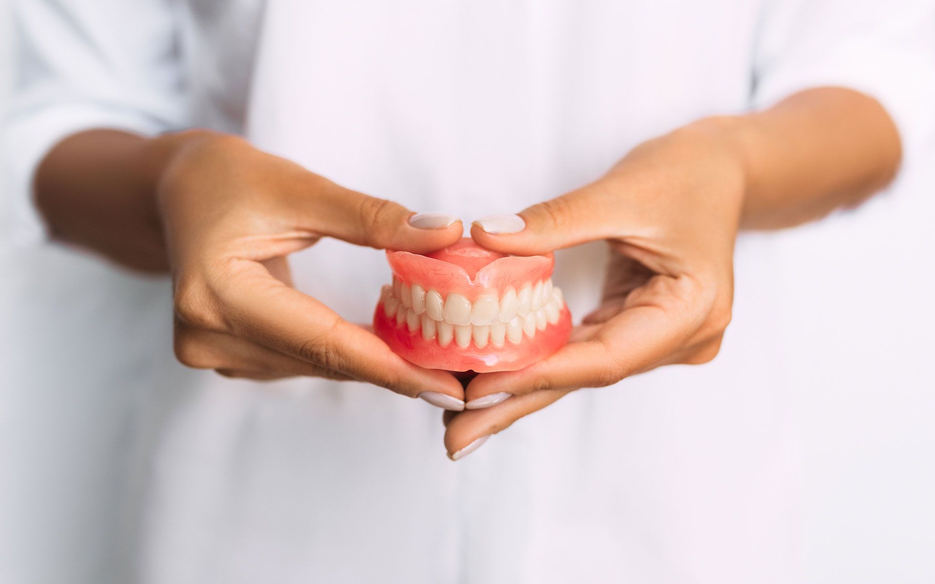 Person holding a set of dentures, showing teeth against a pink gum-like base, white background.