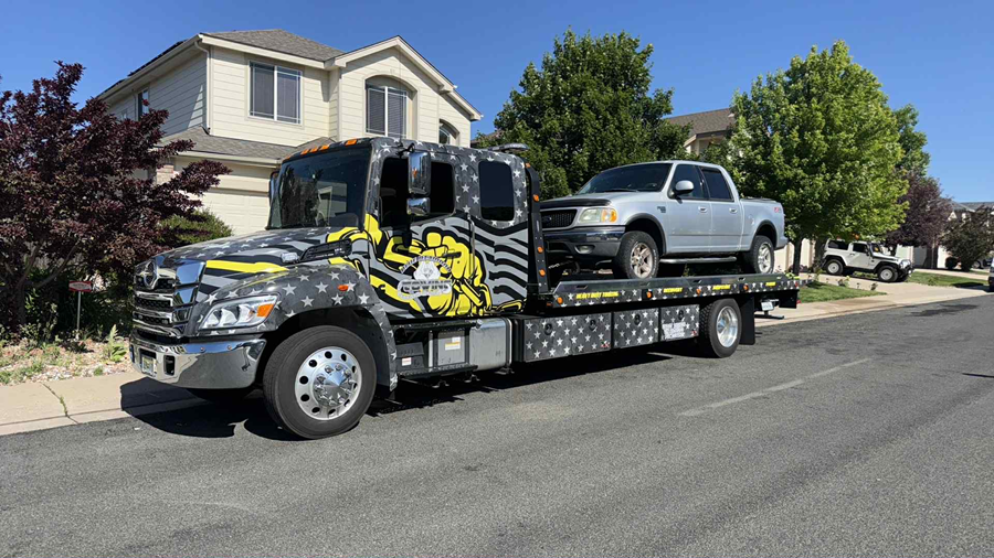 Tow truck with a silver pickup truck on its flatbed, parked on a street in front of a house.