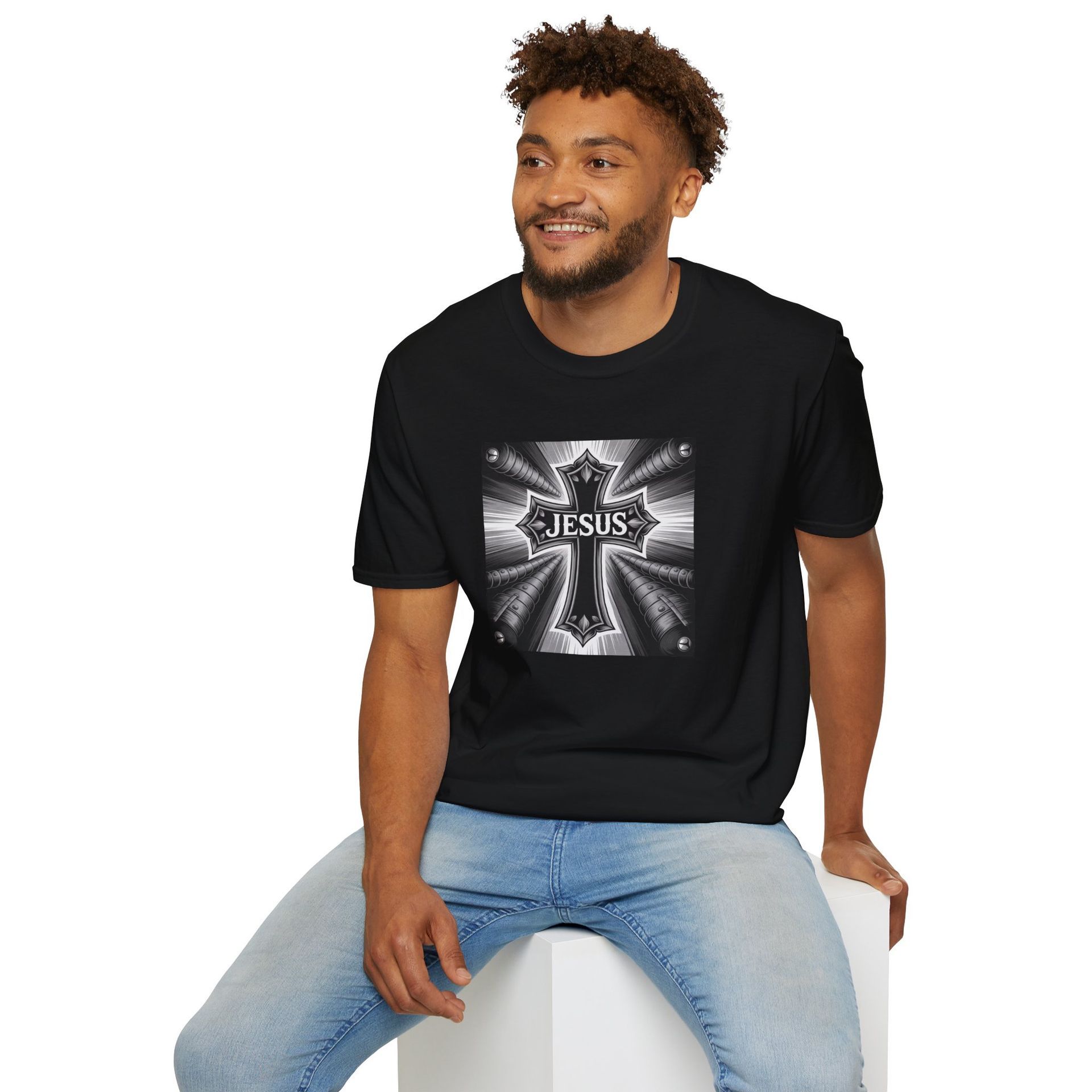 Man wearing black "Jesus" graphic tee, light blue jeans, smiling. Sitting on white cube.